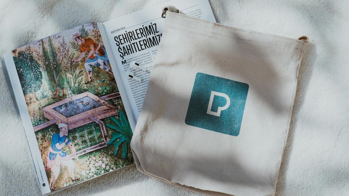 A decorative open book and a tote bag with a logo on a soft textured backdrop.