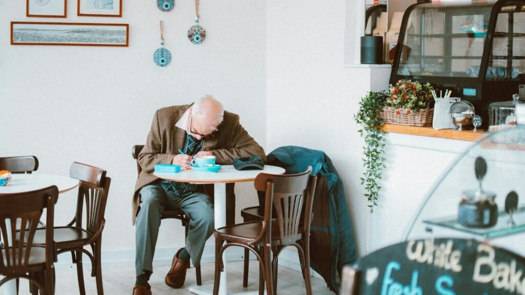 Elderly man writing at a café table, surrounded by cozy decor and ambient lighting.
