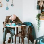 Elderly man writing at a café table, surrounded by cozy decor and ambient lighting.