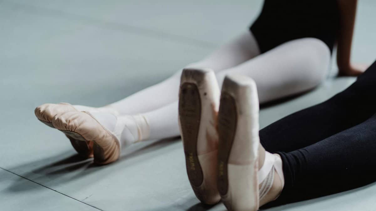 Crop unrecognizable female ballet master with little girl wearing pointes and leggings sitting on floor and doing stretching exercise during training