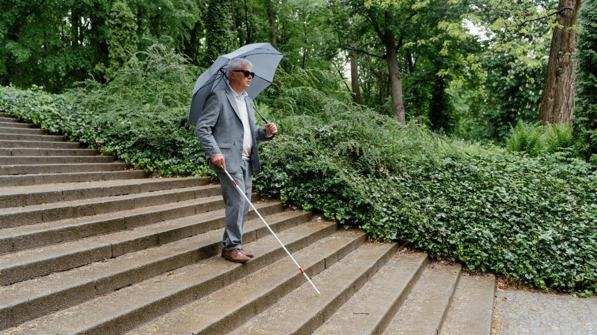 A visually impaired man uses a white cane while walking down outdoor steps with an umbrella.