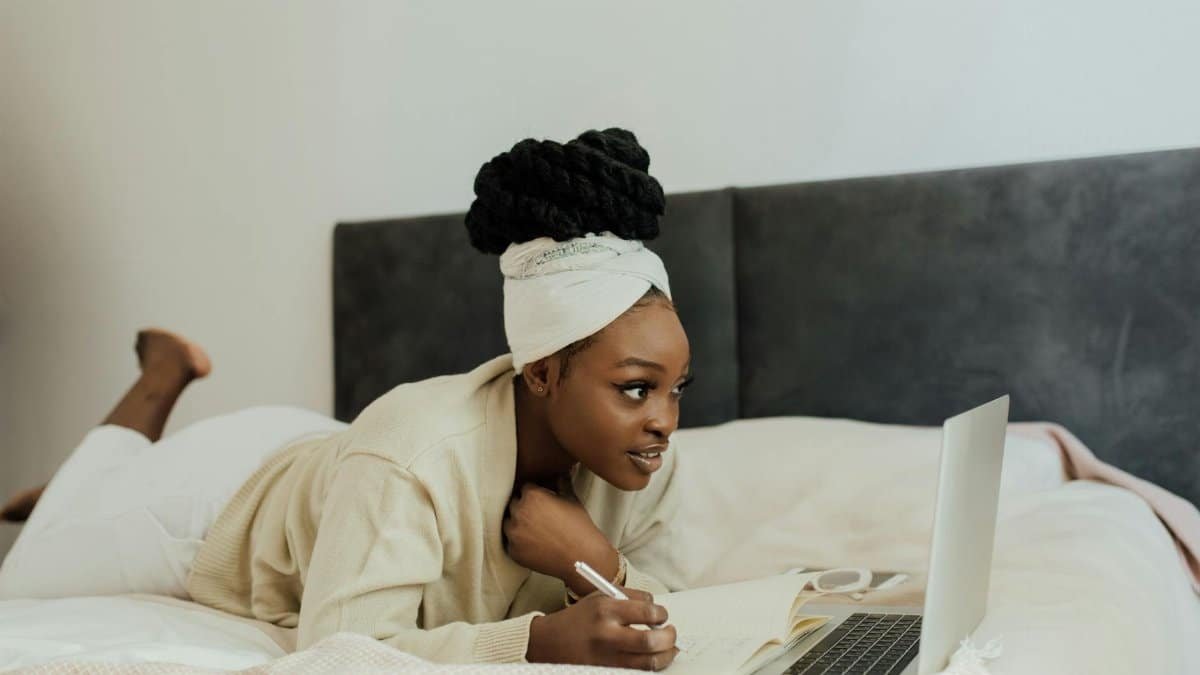 Young woman in headwrap working remotely with laptop and notebook from bed, showcasing a relaxed lifestyle.