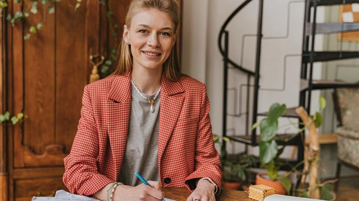 Woman in red blazer smiling while writing at a wooden desk in a cozy home office.