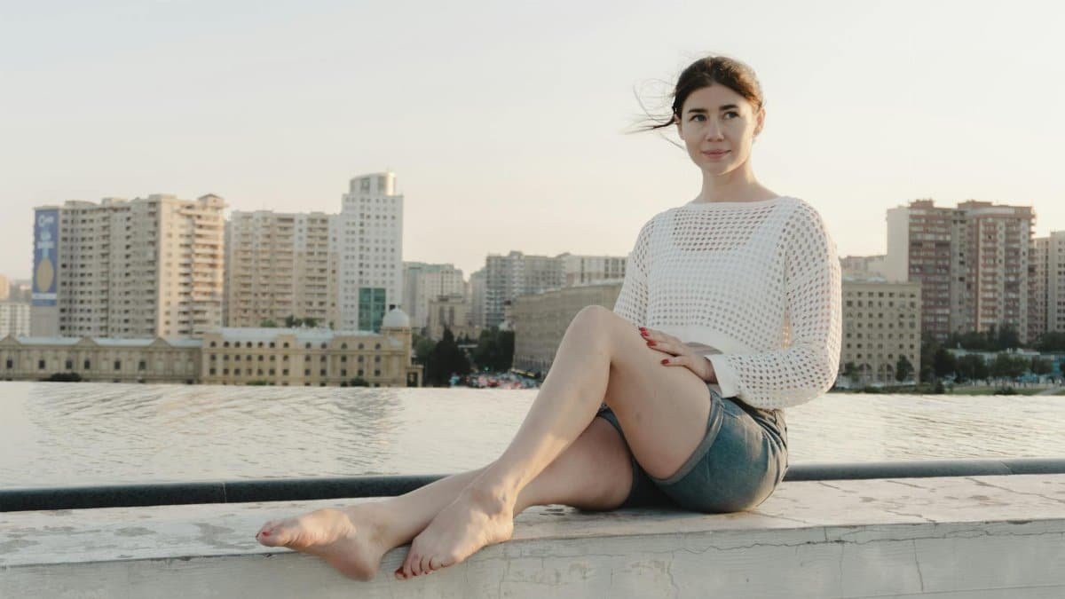 A young woman sitting on a ledge with a city skyline in the background during the day.