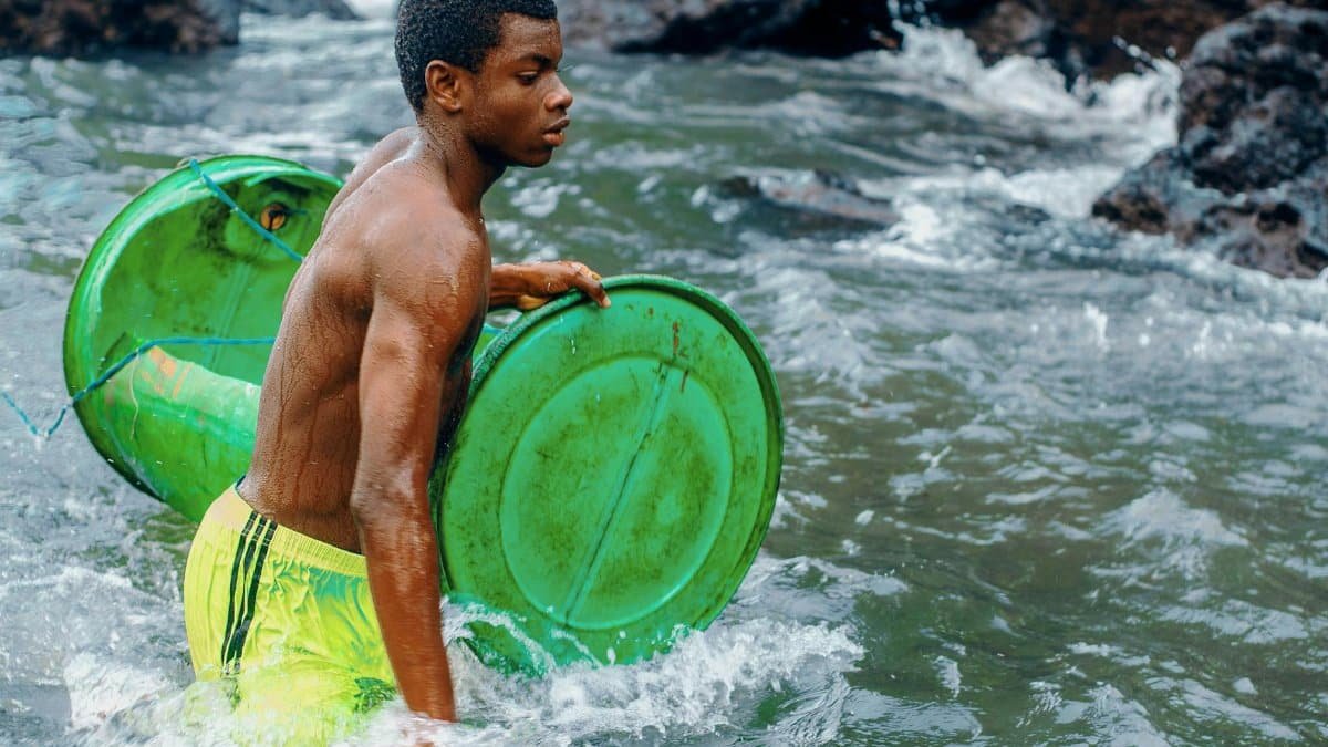 A young man carries green barrels through a river. The scene captures movement and nature.