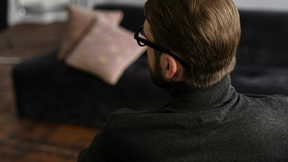 A man with glasses sits contemplatively indoors, suggesting a reflective or therapeutic moment.