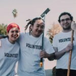 Three diverse volunteers smiling and holding cleaning tools during a community service event.
