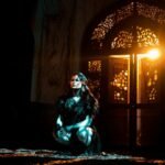 A woman in black attire kneels in dramatic lighting with ornate door backdrop.
