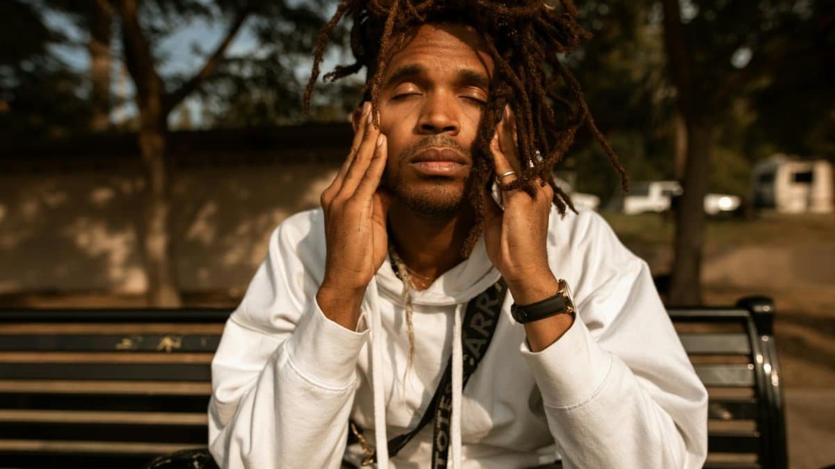 A man with dreadlocks sits on a park bench, contemplating with eyes closed.