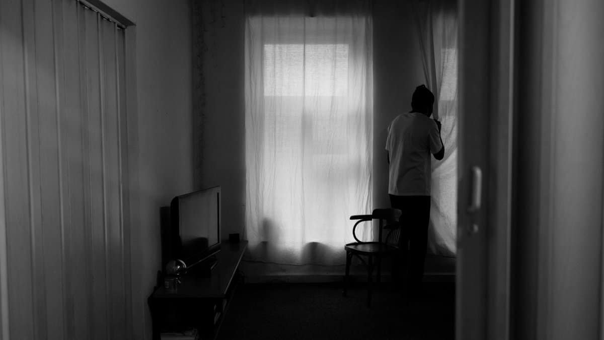 A man stands by a window, creating a silhouette in a dimly lit room, evoking solitude.