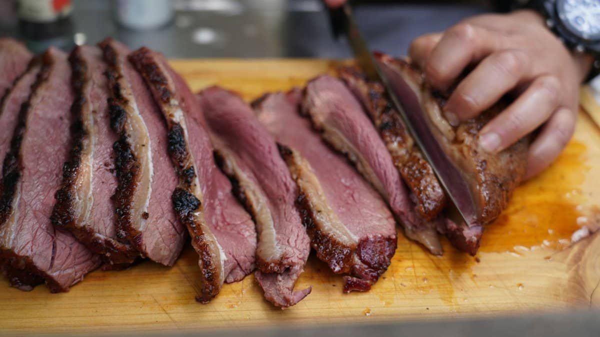 Close-up of a hand slicing juicy, smoked beef brisket on a wooden cutting board, showcasing deliciously cooked meat.