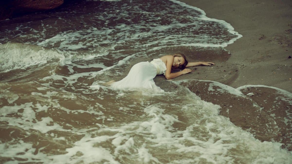 Woman in white dress lying on seashore with gentle waves and sand.