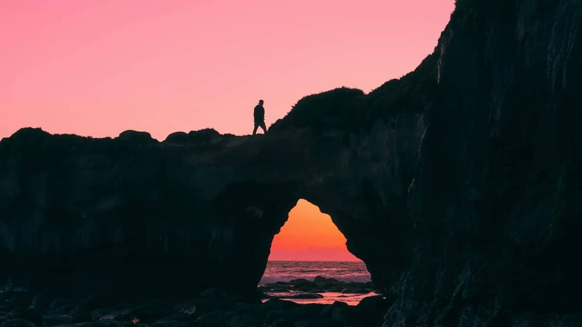 A person silhouetted on a coastal rock arch after sunset with a vibrant sky.