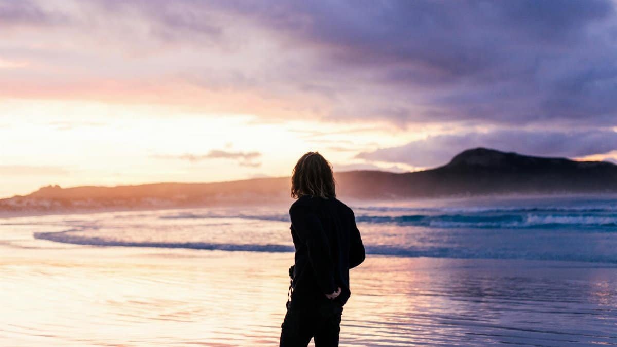 A person walks alone on a calm beach at sunset, surrounded by tranquil sea and moody sky.
