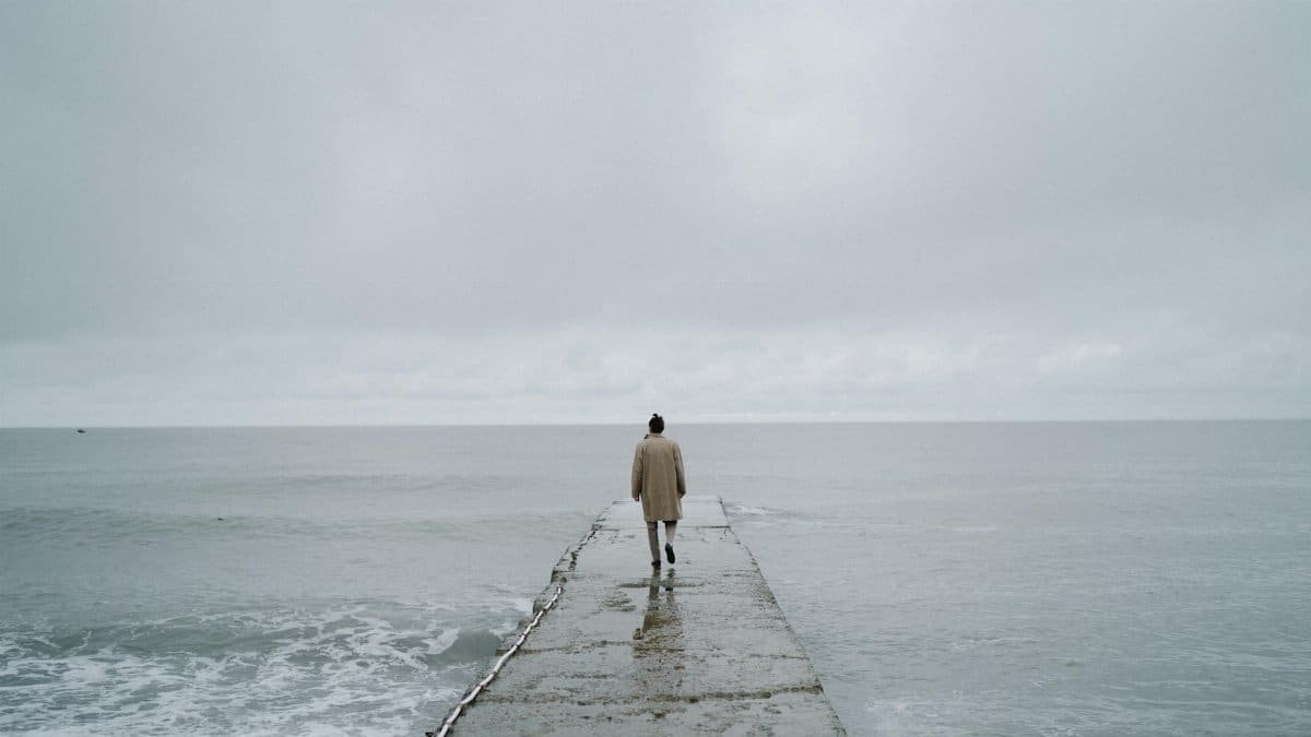 Person in a coat walks alone on a concrete pier towards the vast, open sea under a cloudy sky.