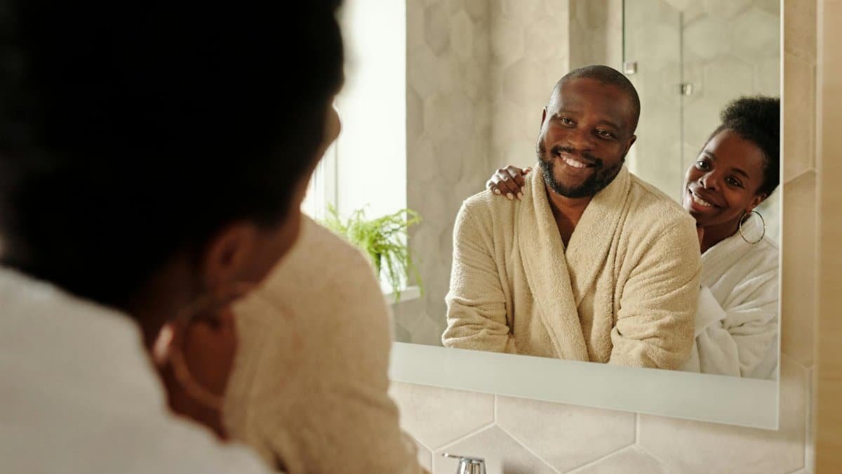 Joyful couple in bathrobes smiling and embracing in bathroom reflection.