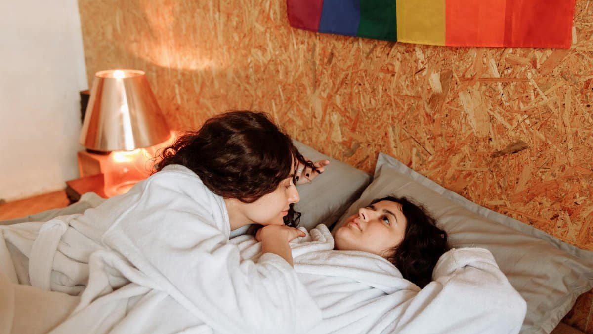 A loving couple embracing in bathrobes under a rainbow flag, creating a warm bedroom atmosphere.