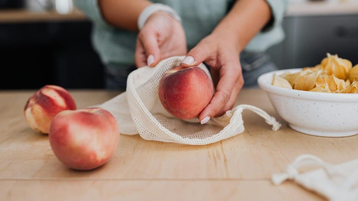 Hands arranging fresh peaches in a reusable net bag on a wooden table for zero waste.