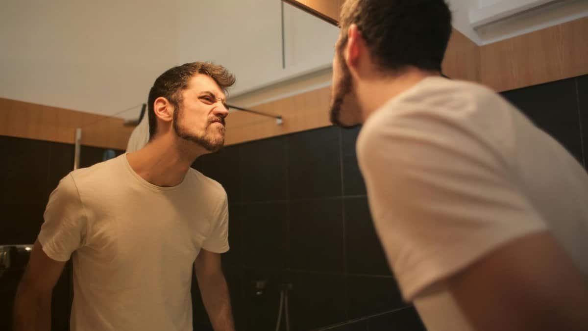 Low angle side view of young bearded male in casual shirt standing in bathroom and looking at with frown mirror in morning