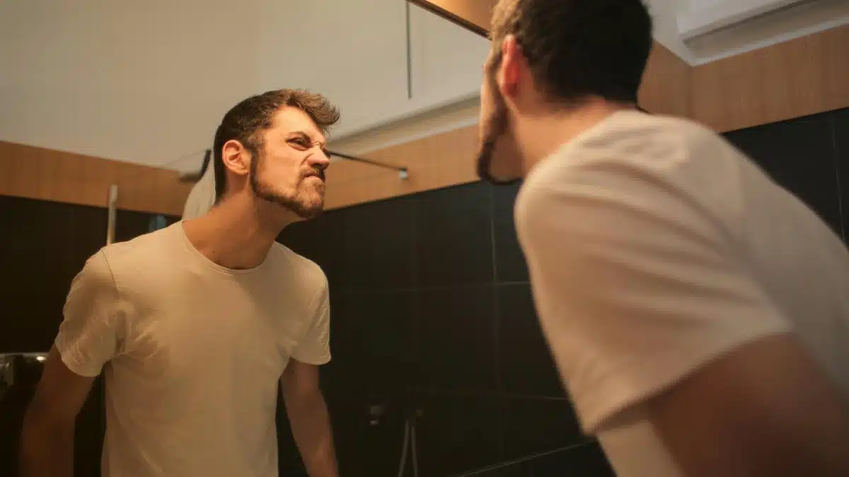 Low angle side view of young bearded male in casual shirt standing in bathroom and looking at with frown mirror in morning