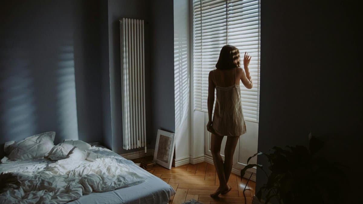 A woman in a bedroom stands by window blinds, casting shadows in soft morning light.