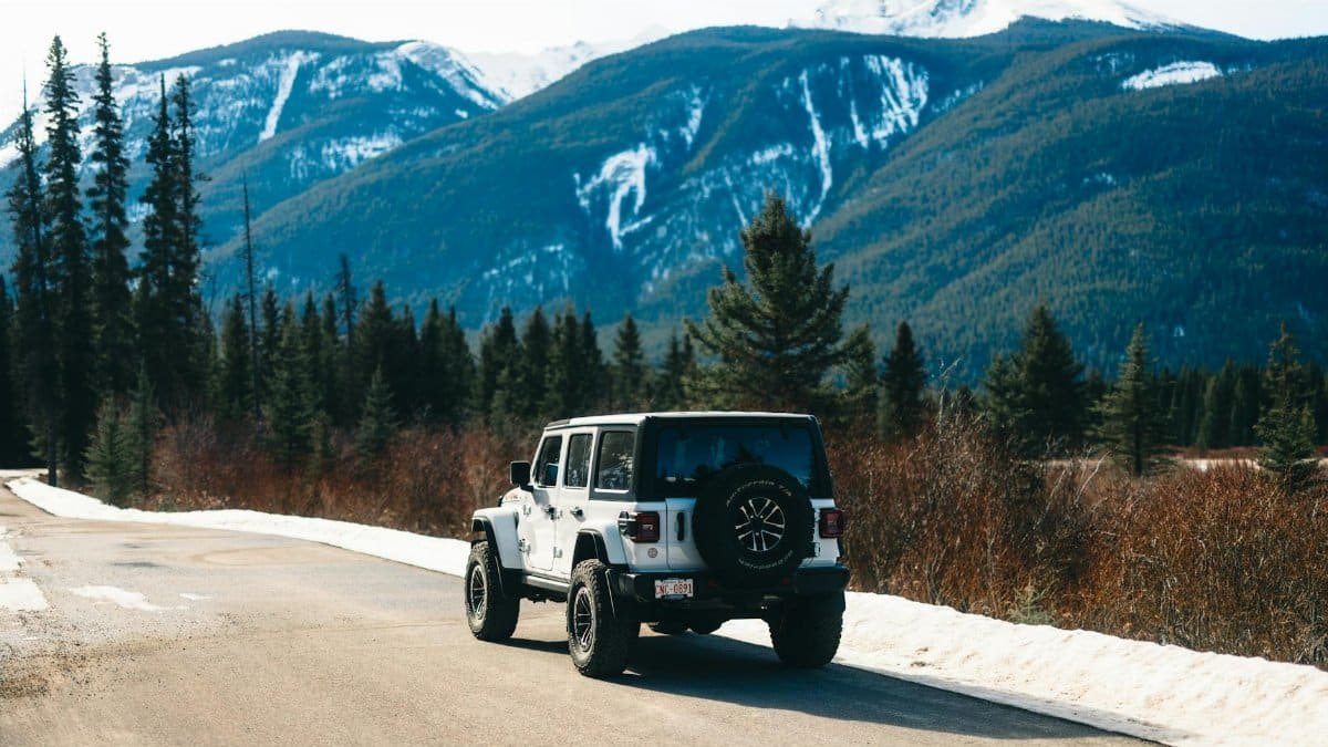 White Jeep on a mountain road in Jasper National Park, Alberta, surrounded by pine trees and snow-capped peaks.