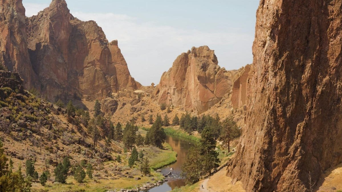 Narrow lake flowing among grassy terrain with trees surrounded with rocky cliffs against cloudy sky in national park on summer day
