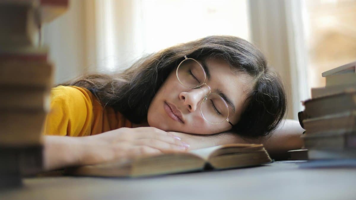 A young woman with glasses naps on a book indoors during the day, capturing a moment of relaxation and fatigue.