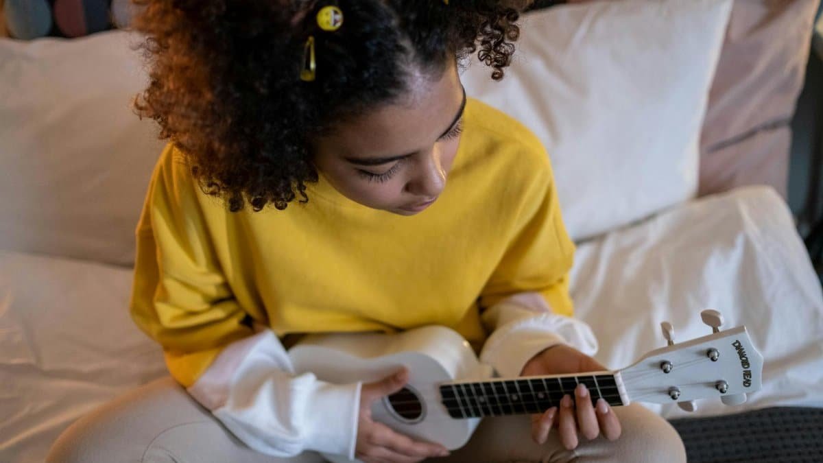 Young girl with curly hair playing a ukulele in a cozy bedroom setting.