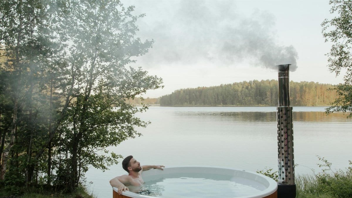 Man enjoying a serene soak in an outdoor hot tub by a peaceful lake surrounded by nature.