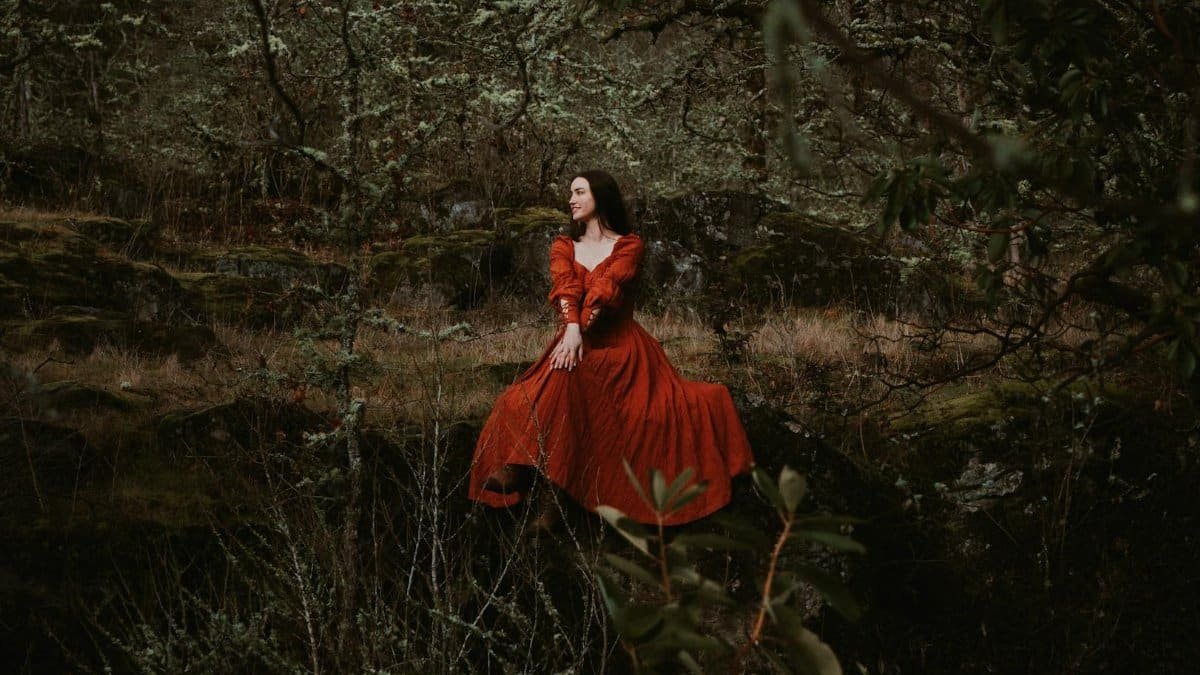 A woman in a red medieval gown poses elegantly in the forest of Portland, Oregon.