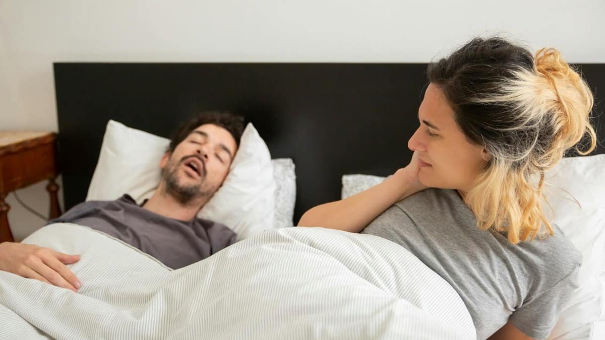 A couple in bed with a man snoring loudly and a woman looking restless on her pillow.