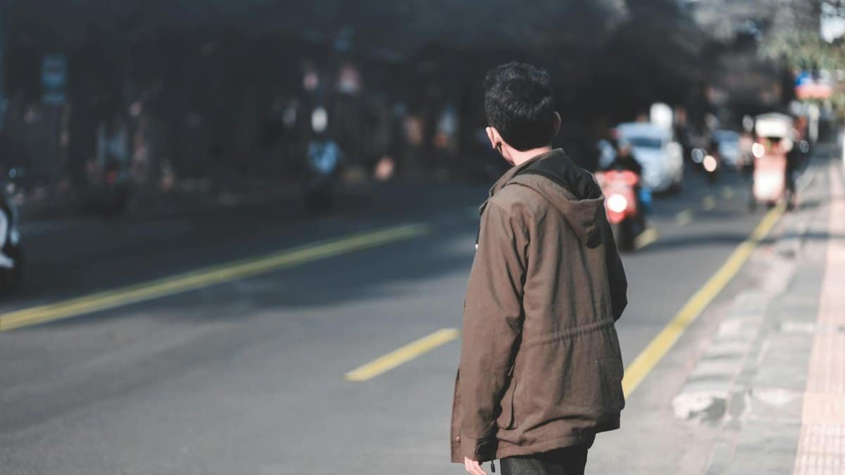 A person in a brown jacket waits on a busy street, observing passing vehicles.