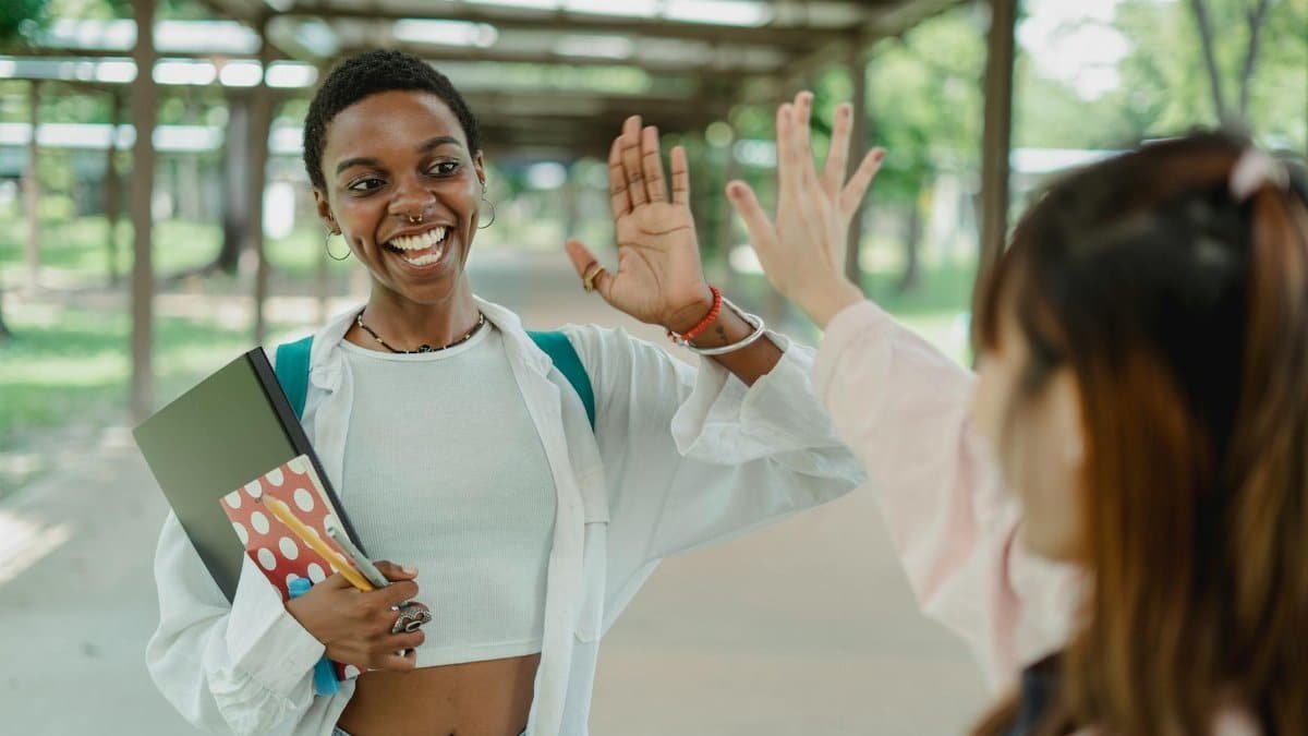 Young content African American female student with workbooks greeting crop anonymous partner on roofed pavement while looking at each other