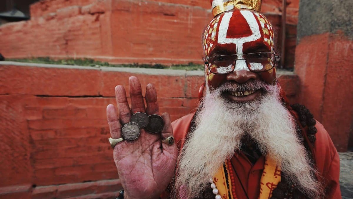 A joyful Indian sadhu with traditional attire, showcasing coins and a vibrant smile in an outdoor setting.