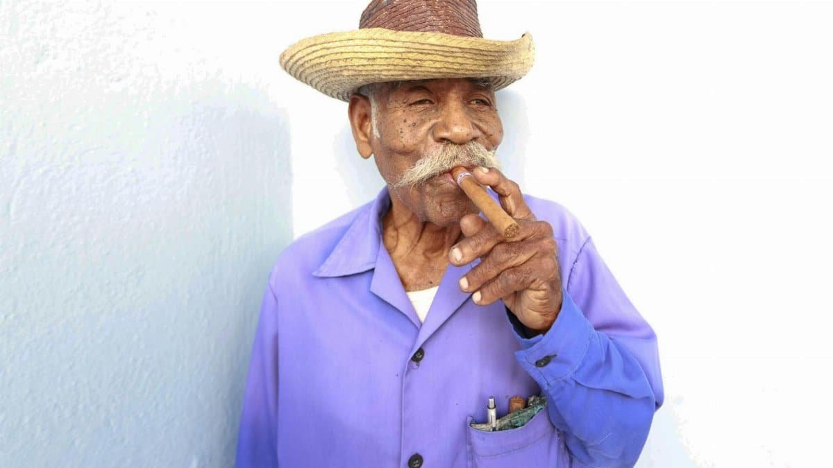 Portrait of an elderly man wearing a straw hat and blue shirt, smoking a cigar against a white background.