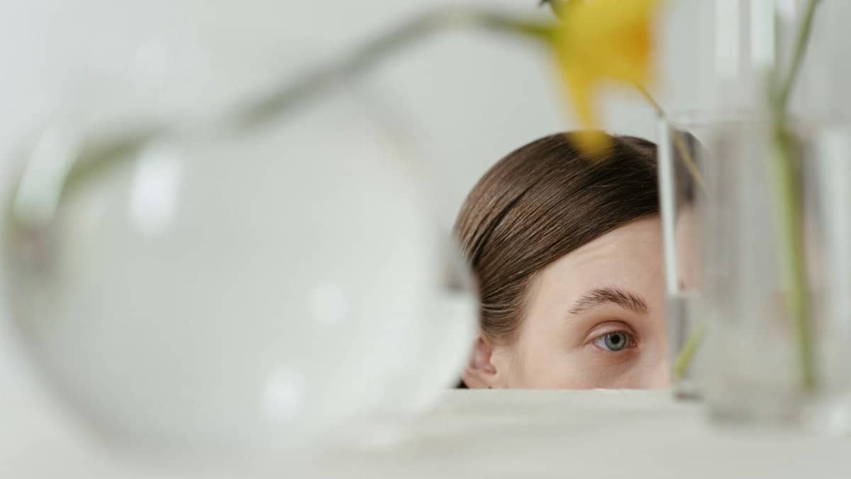 Close-up of a woman's eye peering from behind a glass vase holding flowers.