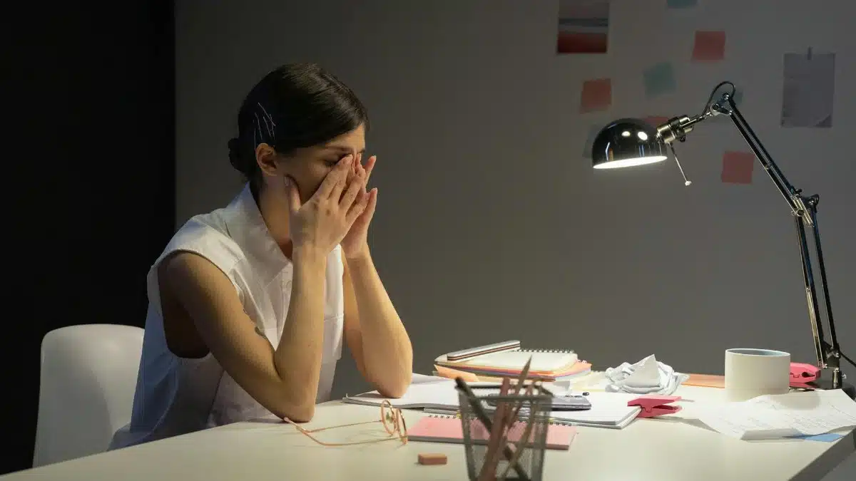 A stressed businesswoman rubs her eyes at a cluttered desk, overwhelmed by work.
