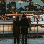 Couple embraces in winter attire overlooking ice rink in festive Toronto setting.