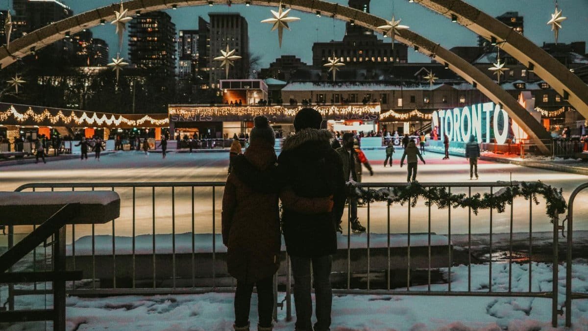 Couple embraces in winter attire overlooking ice rink in festive Toronto setting.