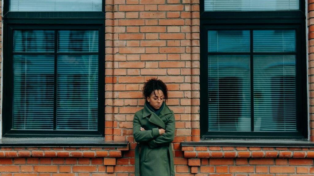 A woman in a green coat leans against a brick wall, appearing thoughtful and introspective.