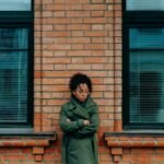 A woman in a green coat leans against a brick wall, appearing thoughtful and introspective.