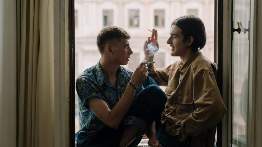 Two men sitting on a windowsill sharing a cigarette, capturing a moment of connection.