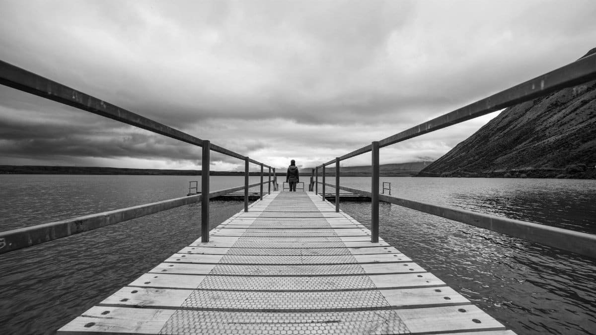 Black and white photo of a person on a pier, capturing solitude amidst a dramatic lakeside setting.