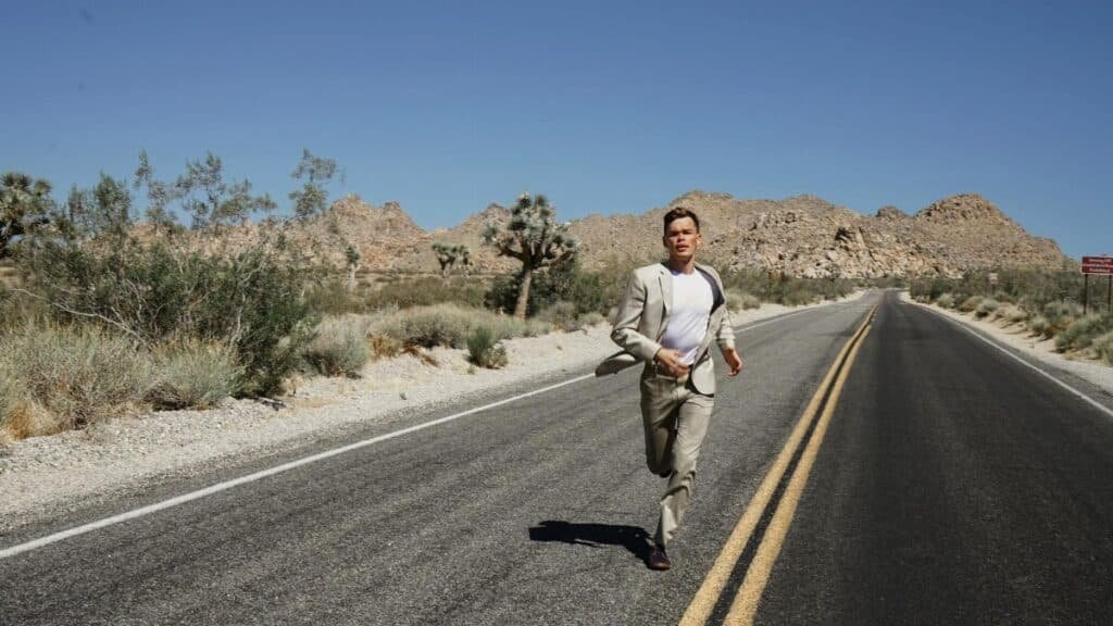 A man in a suit running on an empty desert road surrounded by mountains and dry vegetation under a clear blue sky.