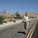 A man in a suit running on an empty desert road surrounded by mountains and dry vegetation under a clear blue sky.