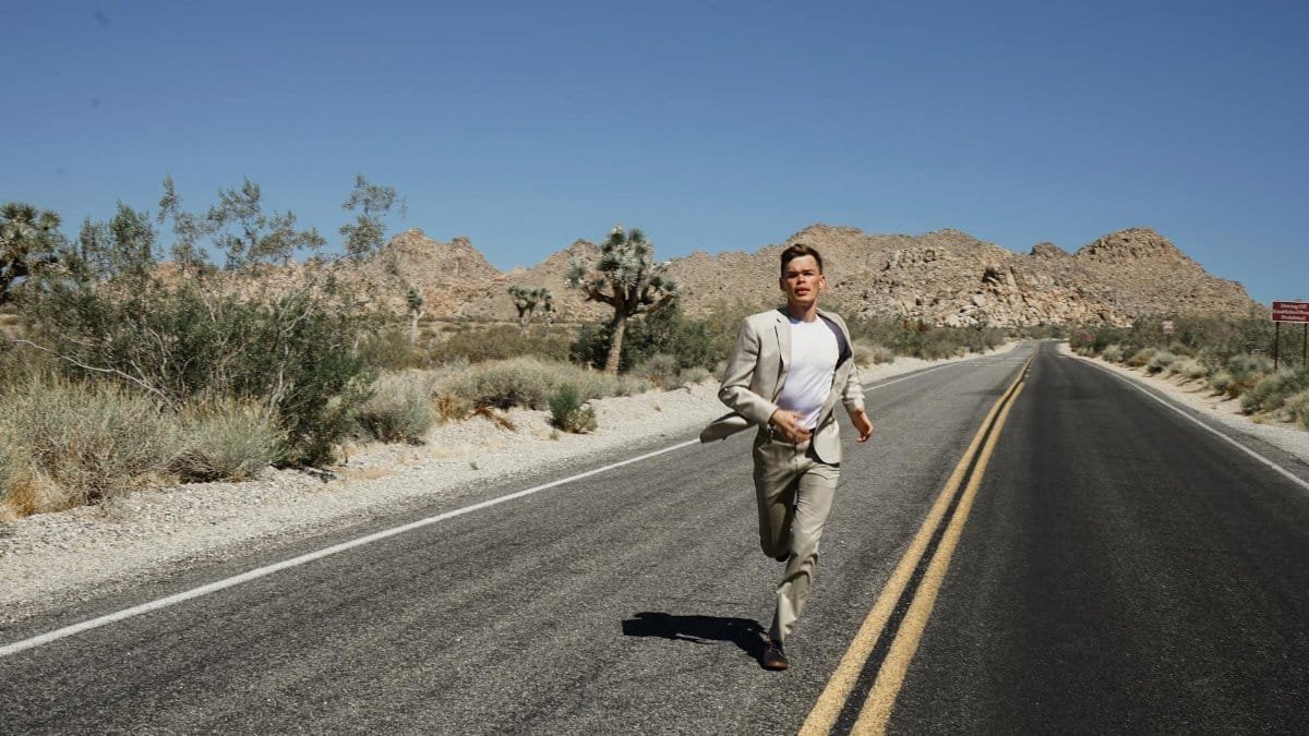 A man in a suit running on an empty desert road surrounded by mountains and dry vegetation under a clear blue sky.