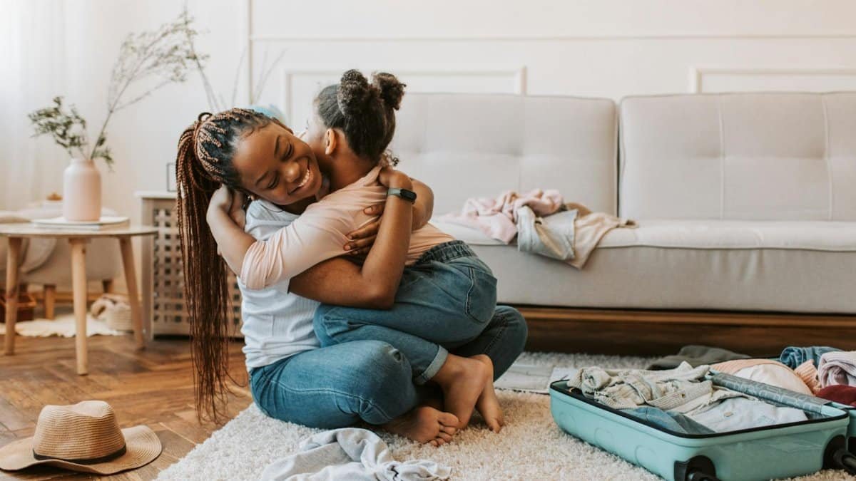 A mother and daughter share a loving hug while preparing for a trip with an open suitcase at home.