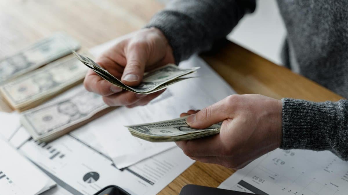 Person counting dollar bills on a desk with financial documents and a calculator in the background.