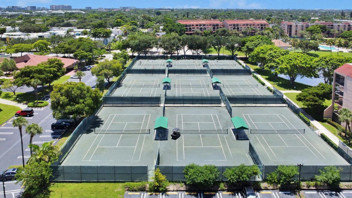 Aerial shot of tennis courts surrounded by trees and buildings in a sunny urban setting.